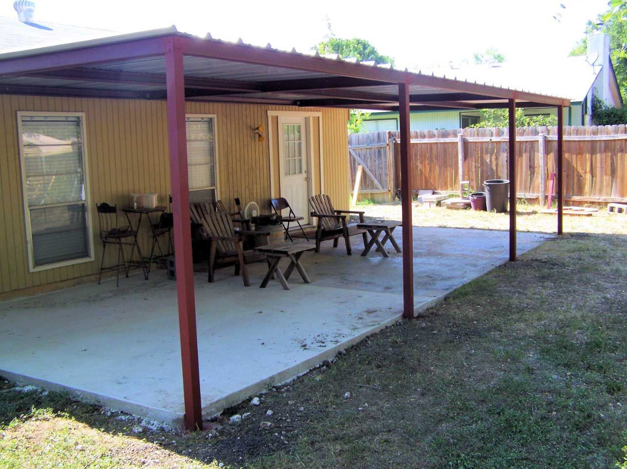 A concrete patio with a red-framed metal roof, outdoor furniture, and a grassy yard.