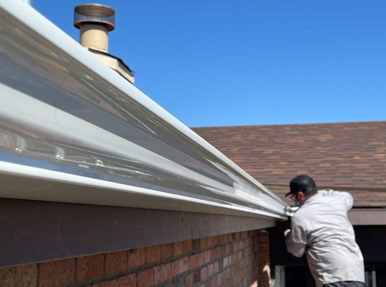 Man installing a white gutter on a brick house roof under a clear blue sky.