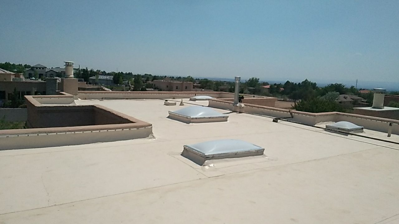 Overhead view of a flat, light-colored roof with skylights, chimneys, and surrounding buildings under a clear sky.