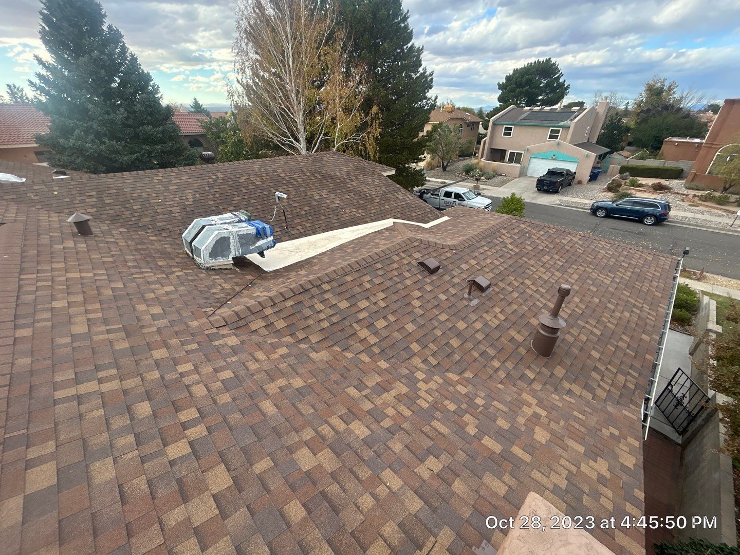 Brown shingle roof with an HVAC unit on top, houses in background, trees, and blue sky.