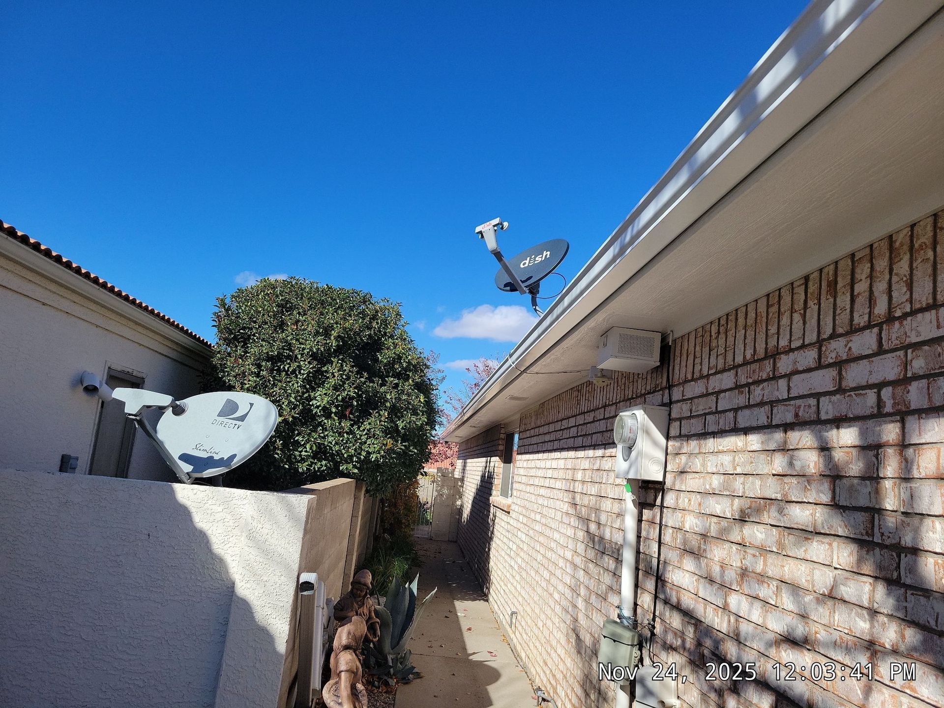 Two satellite dishes mounted on buildings in a narrow alleyway against a bright blue sky.