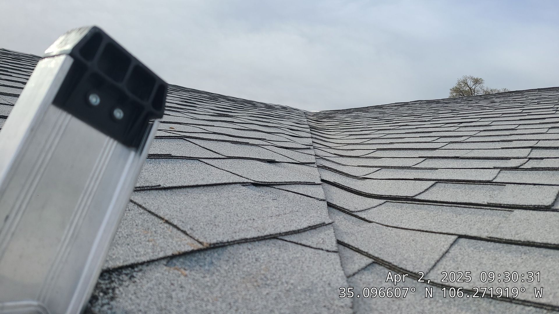 Ladder on a gray shingled roof, angled towards a roof valley, cloudy sky.