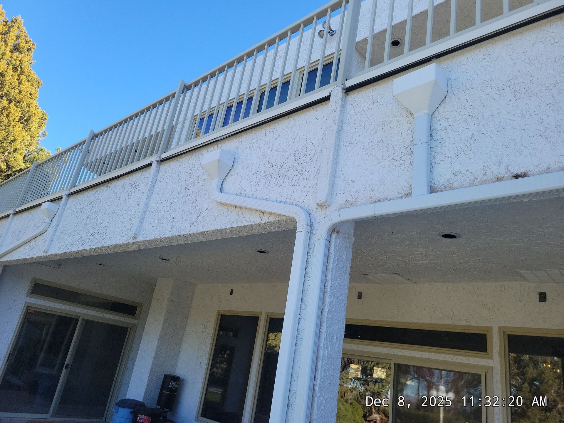 White building with balcony and railings. Blue sky overhead. Downspouts and windows visible.