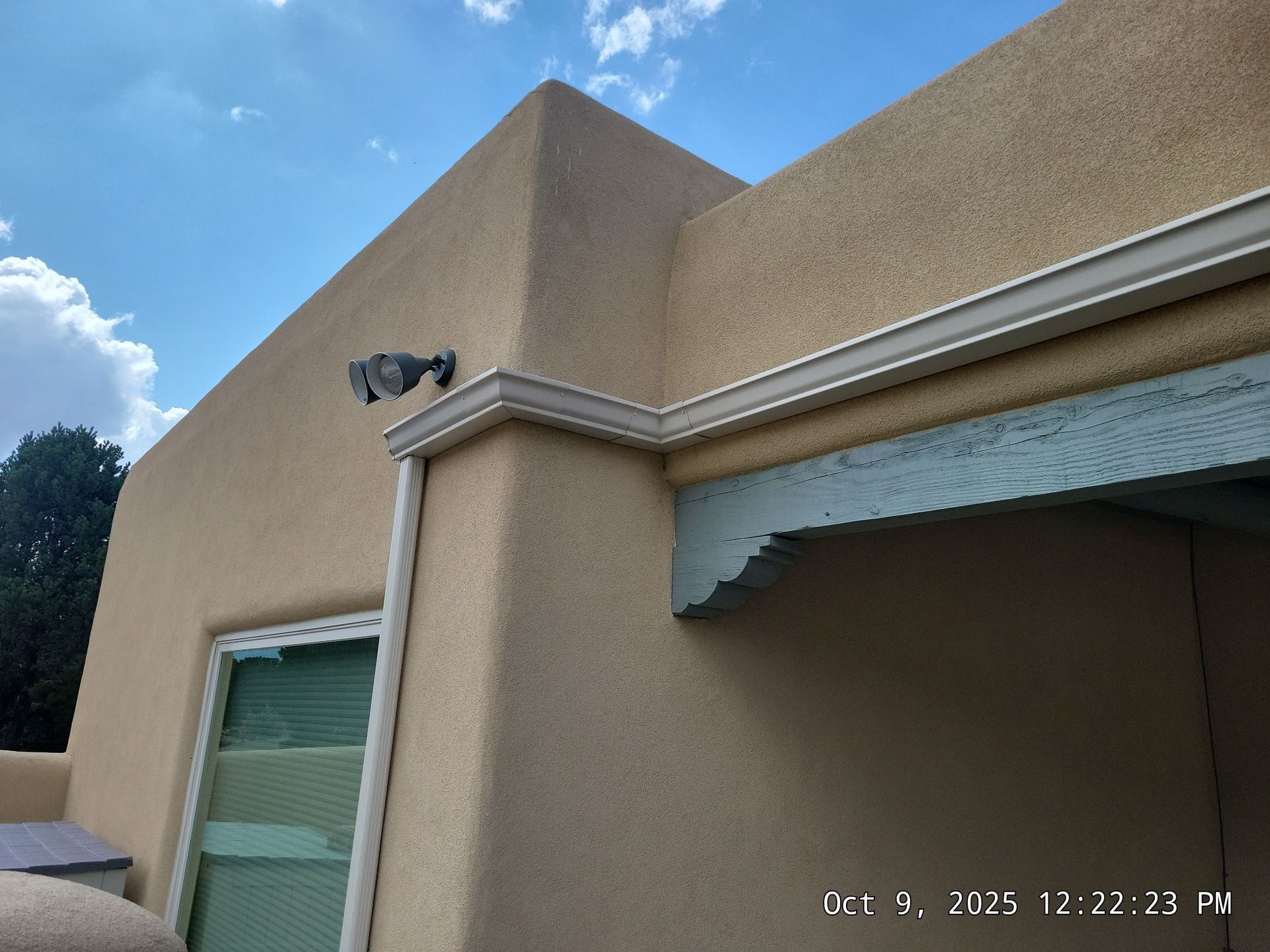 Tan stucco building corner with a security camera, white trim, and a window against a blue sky.
