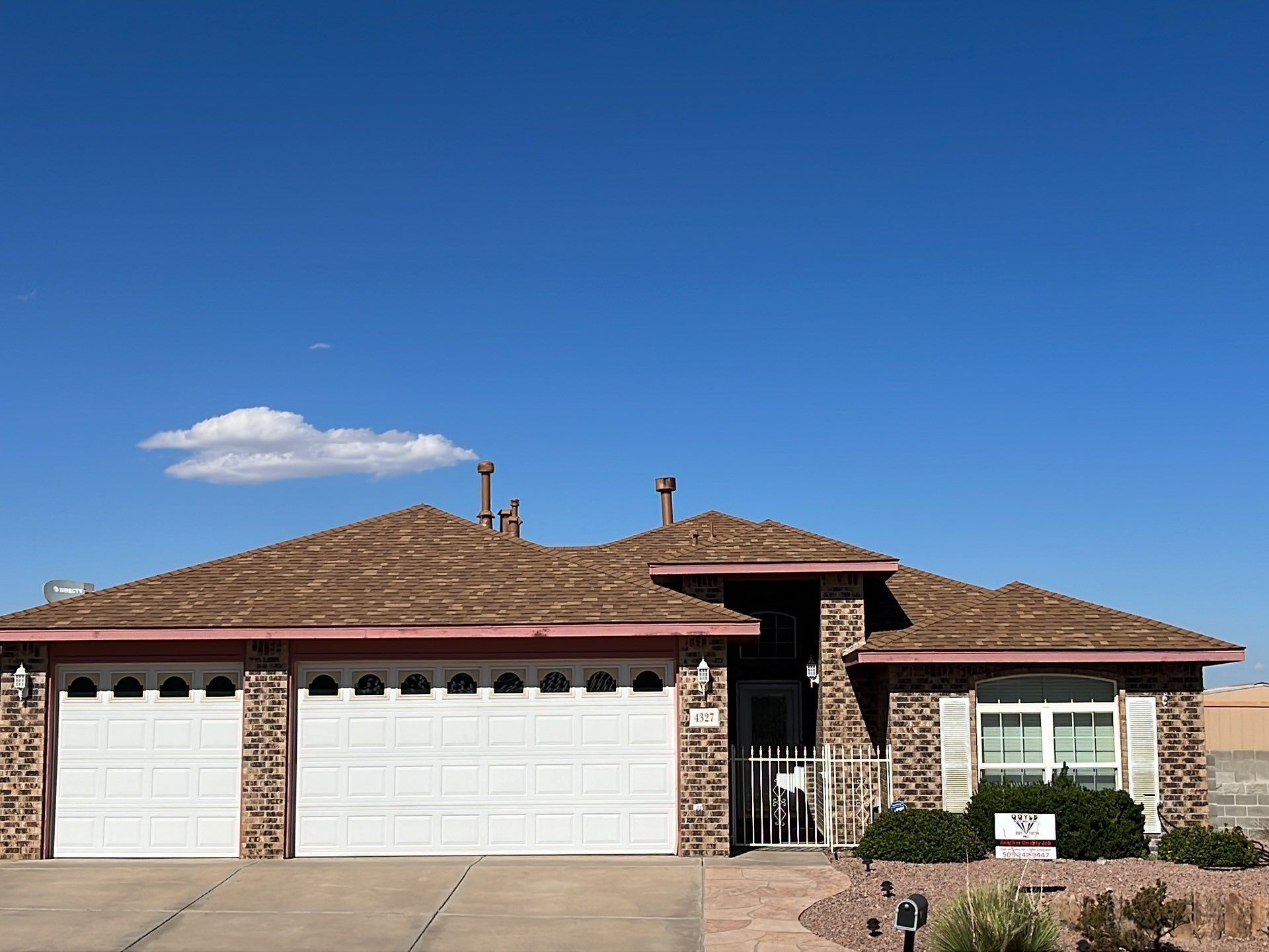 House with a three-car garage, brick facade, and tan roof against a blue sky with a cloud.