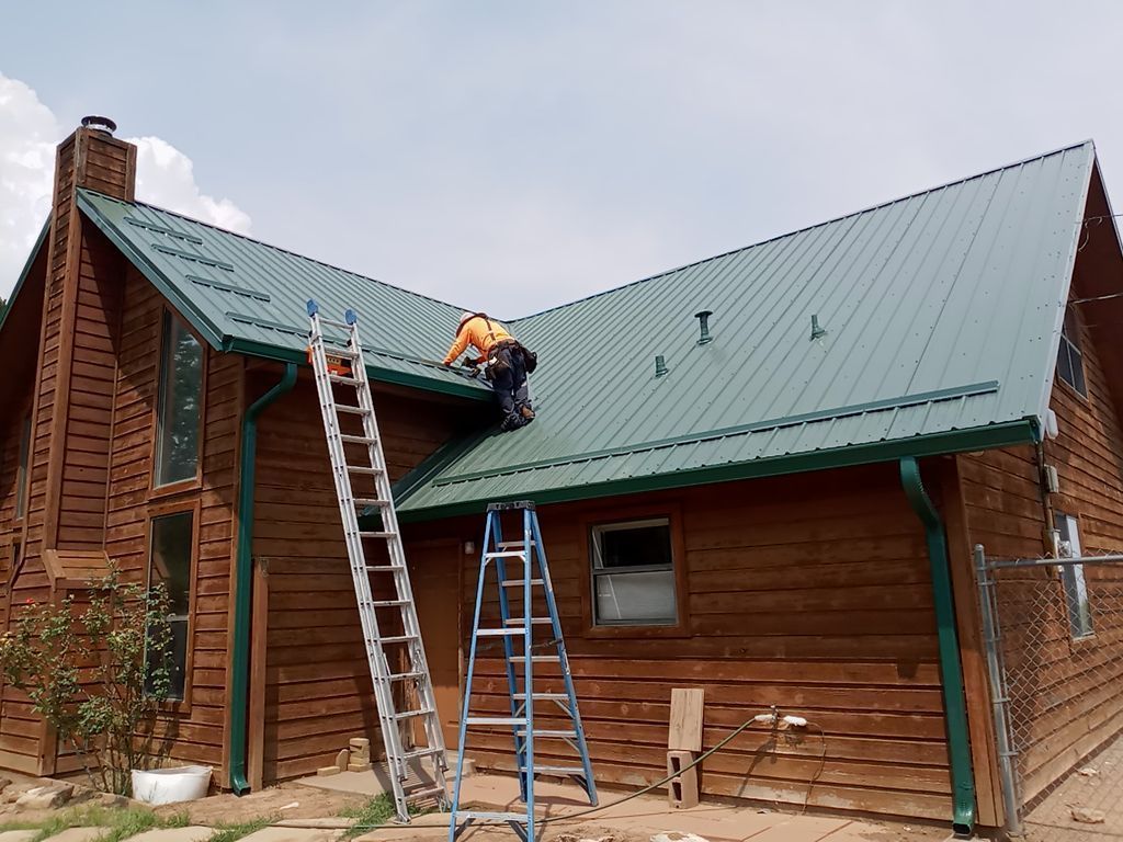 Two workers on a green metal roof of a log cabin, using ladders.