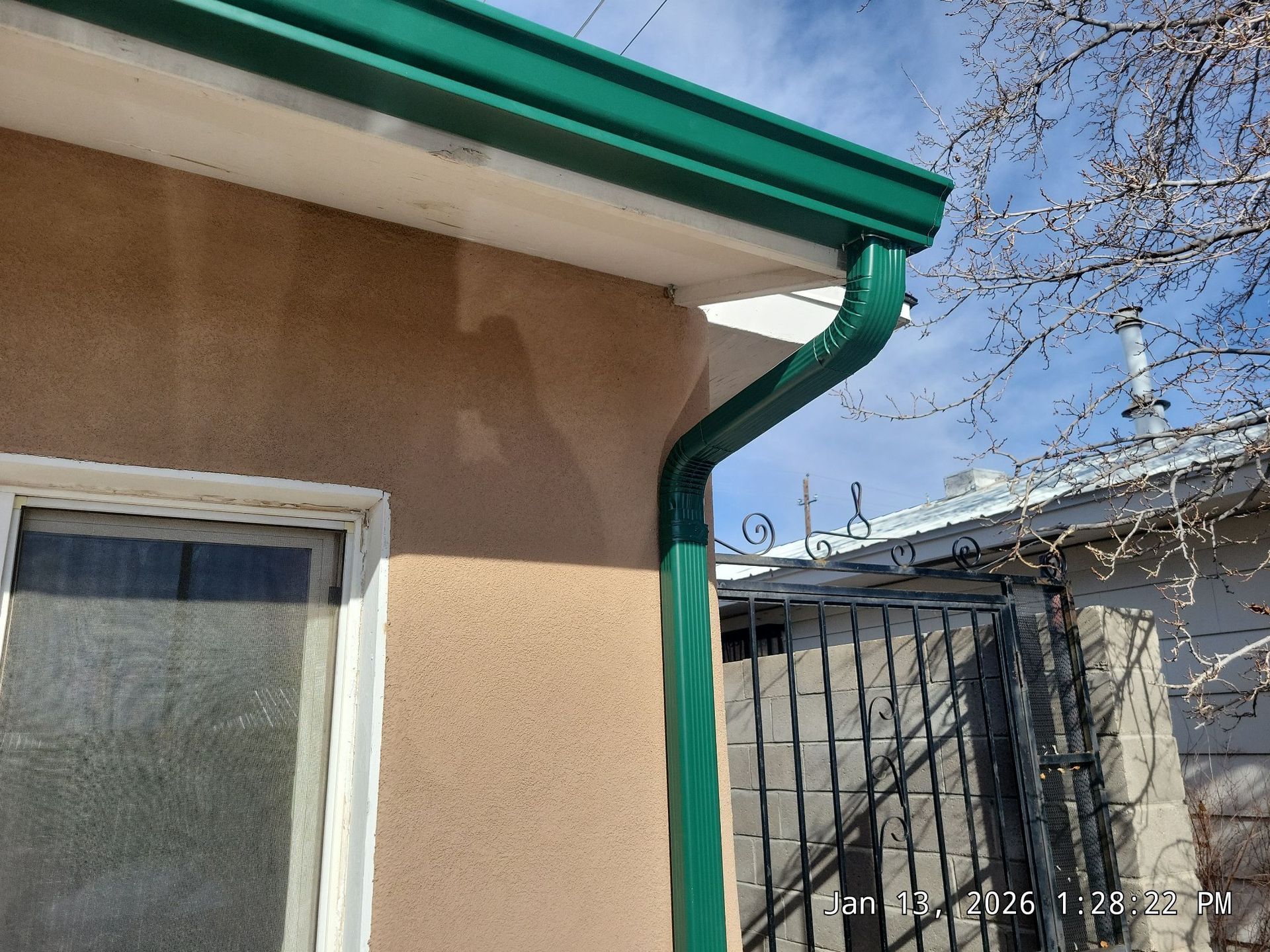 Green rain gutter on a tan stucco house with a window; clear sky background.