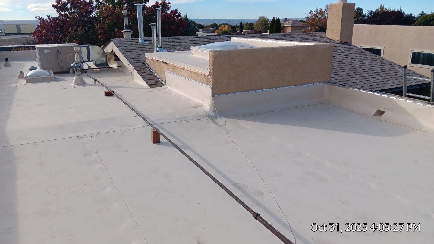 A white flat roof with vents, a chimney, and a building in the background against a blue sky.