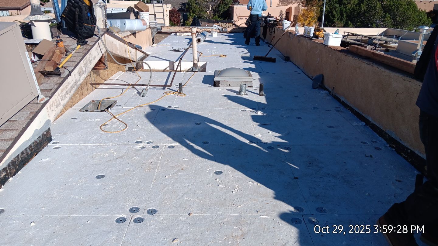 A flat roof with workers. Gray and white surfaces, shadows. Tools and materials scattered. Clear, sunny day.