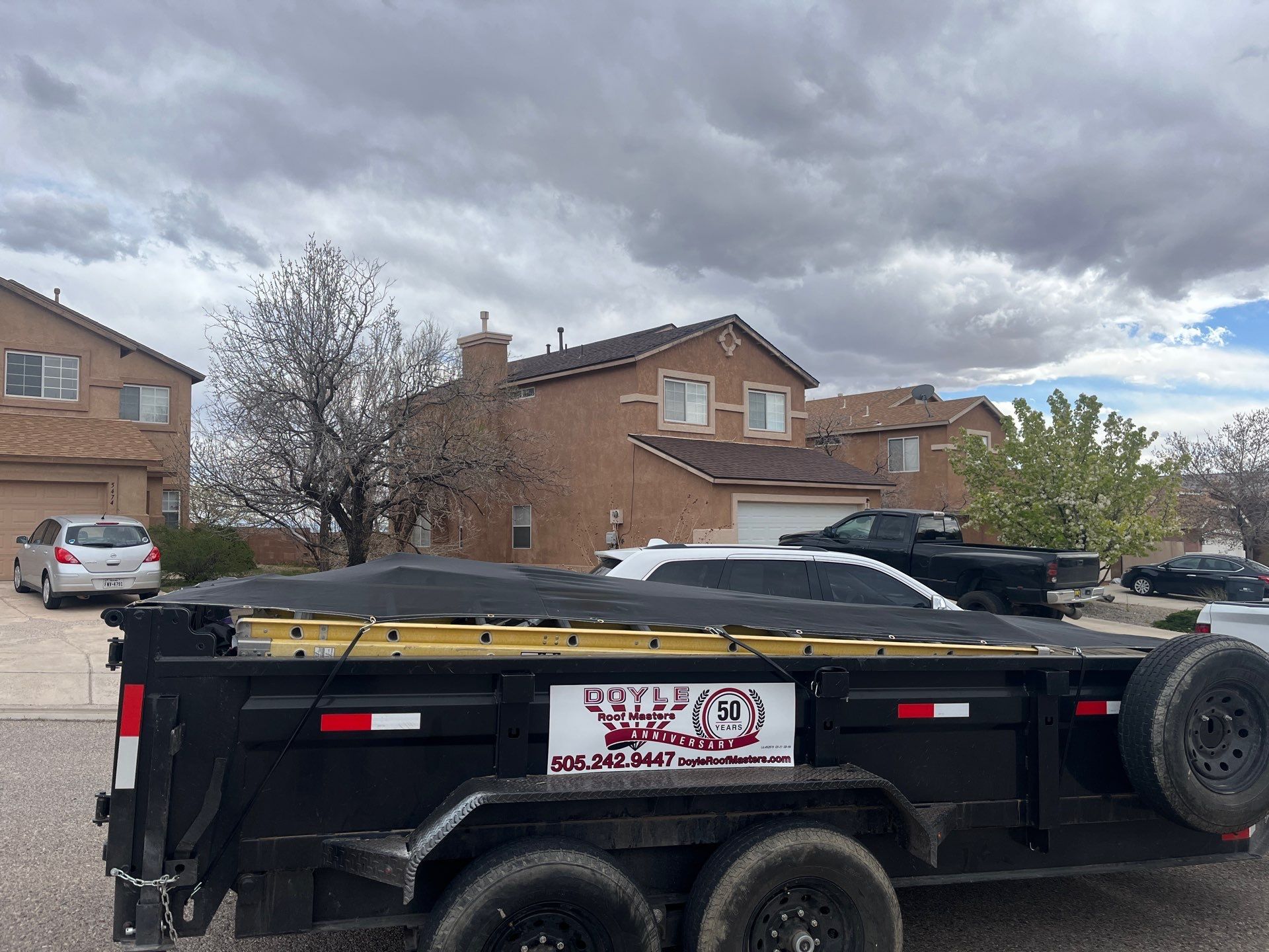 Black utility trailer with a tarp cover, parked in front of suburban houses on a cloudy day.