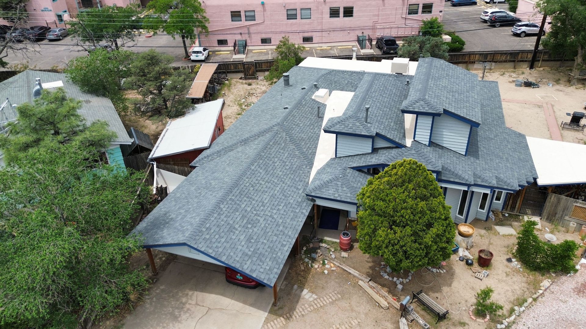 Aerial view of a blue-roofed house with carport, surrounded by trees and a pink building in the background.