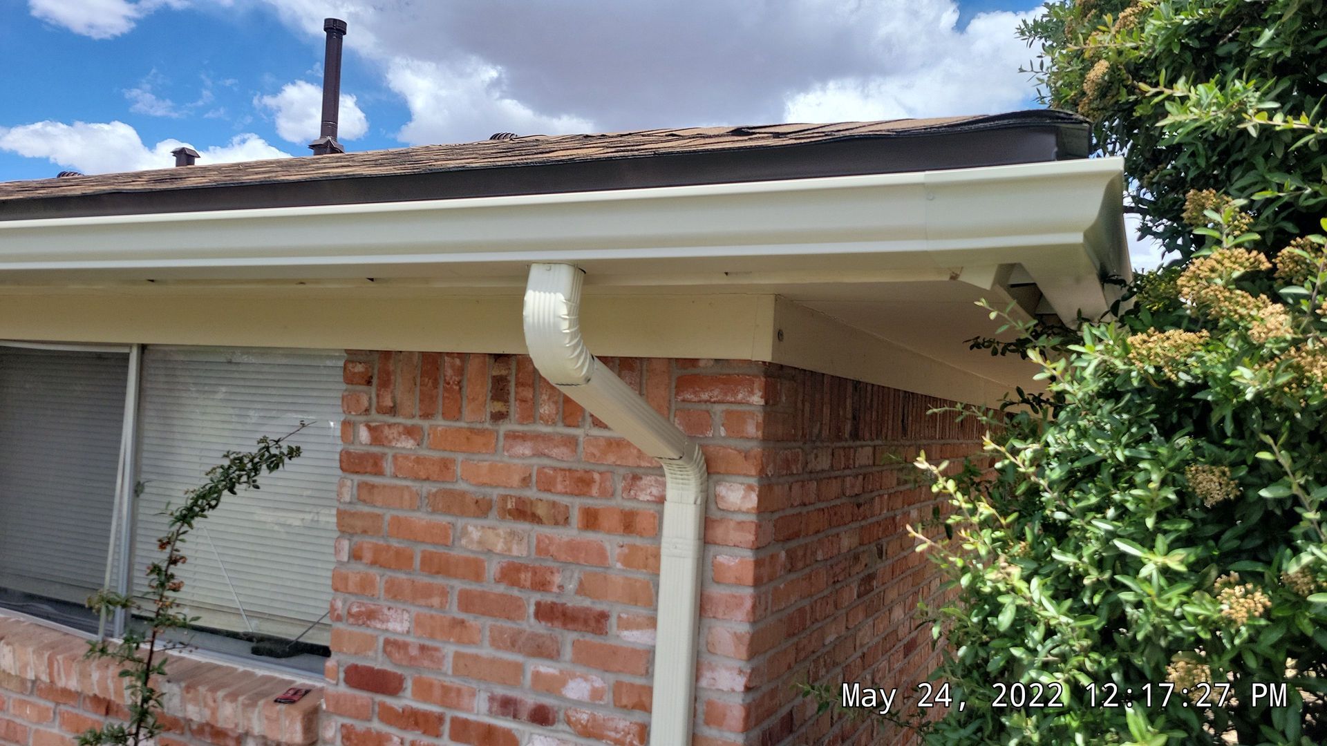 Beige gutters and downspout on a brick building with a leafy bush in the right corner.