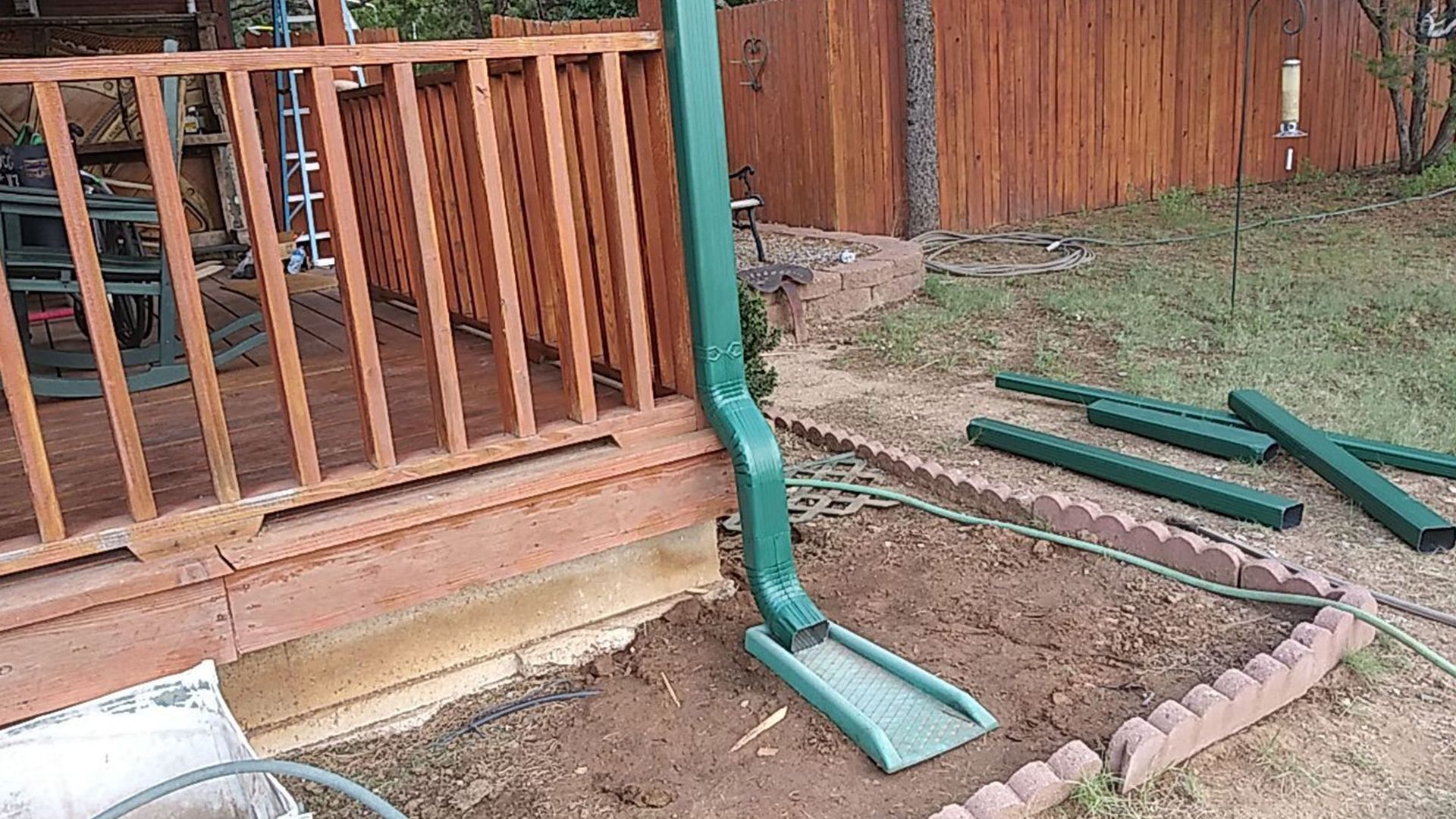 Green downspout next to a wooden deck, with scattered green gutter components on the ground.