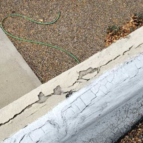 Cracked concrete wall edge with peeling paint, gravel ground, and green hose in the background.