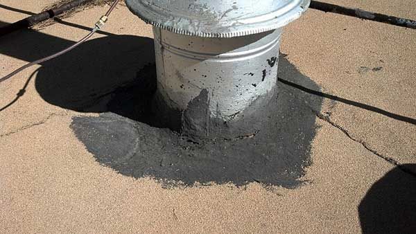 Metal vent pipe sealed with black sealant on a tan rooftop.