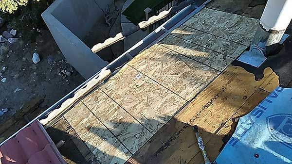 Rooftop view of OSB boards being installed. A person's legs are visible. Blue underlayment present. Sunny.