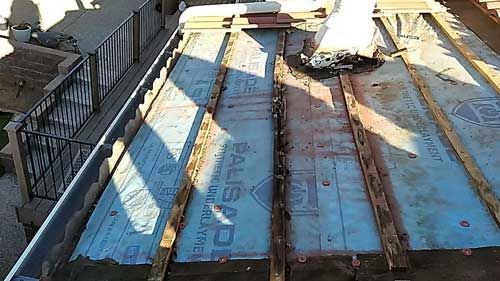 Roof with exposed underlayment and support beams, near a gutter and railing.