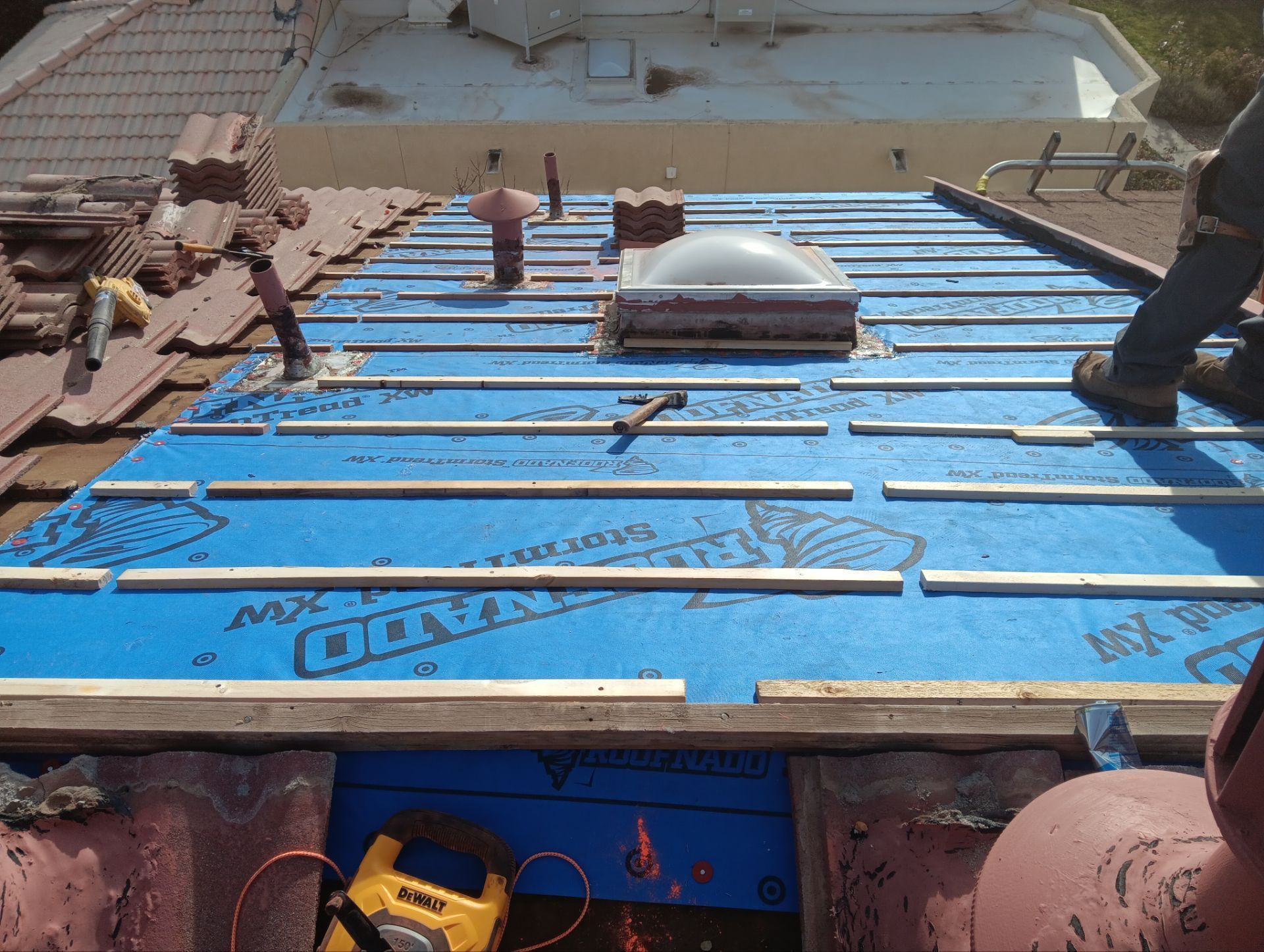 Roof under construction; blue underlayment with metal flashing strips, skylight, tiles, and tools visible.