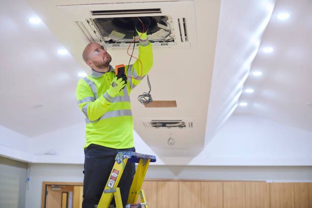 Technician on ladder repairing ceiling air vent with exposed wiring in a room