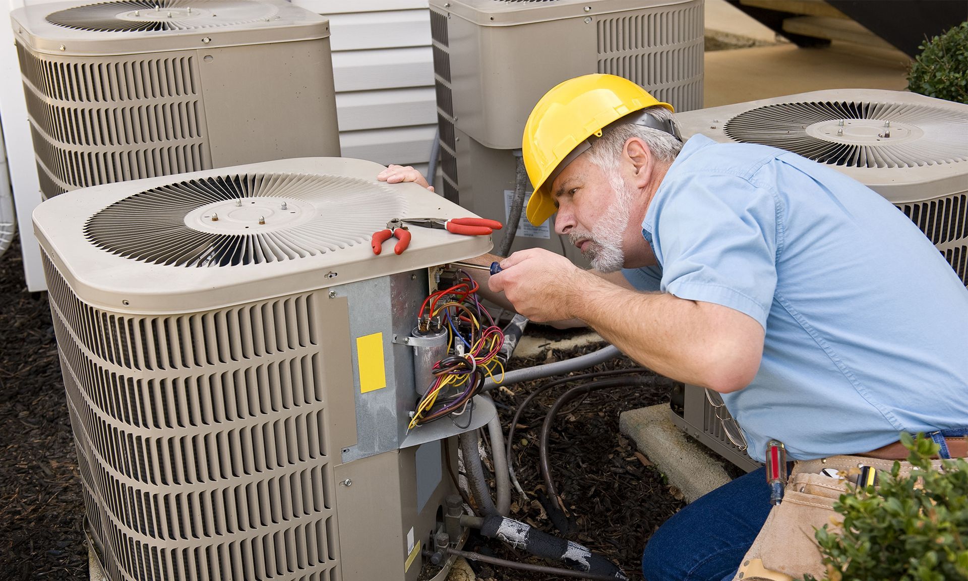 Technician in hard hat repairing outdoor HVAC units with exposed wiring.