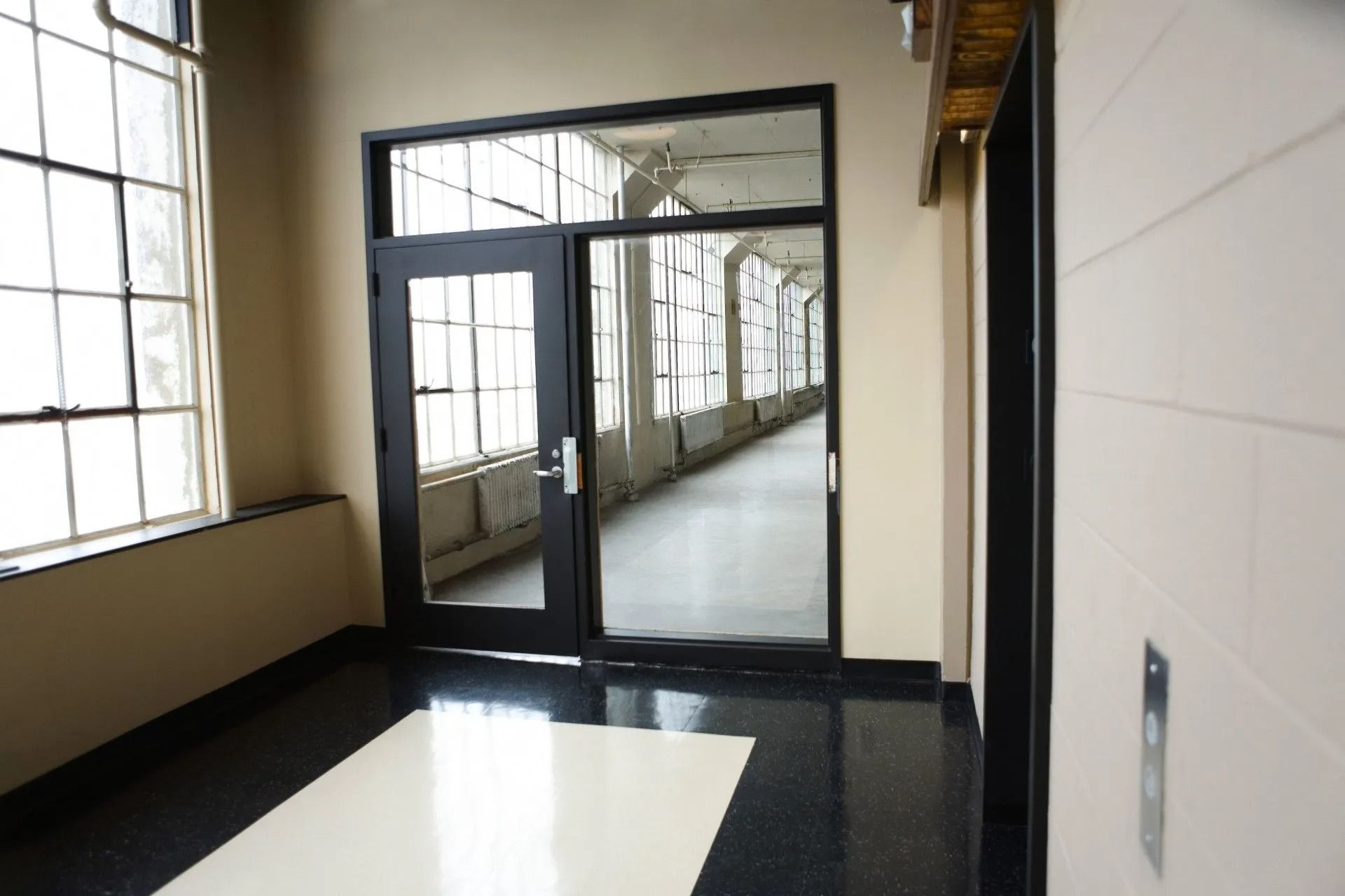 Hallway with a glossy black floor, a glass door leading to a bright corridor, and windows on the left.