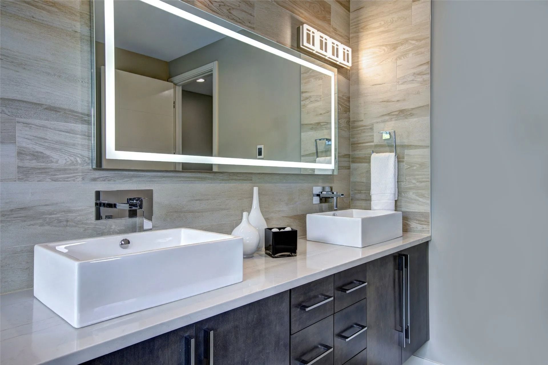 Modern bathroom with a double vanity, featuring rectangular sinks, a large backlit mirror, and light gray tile walls.