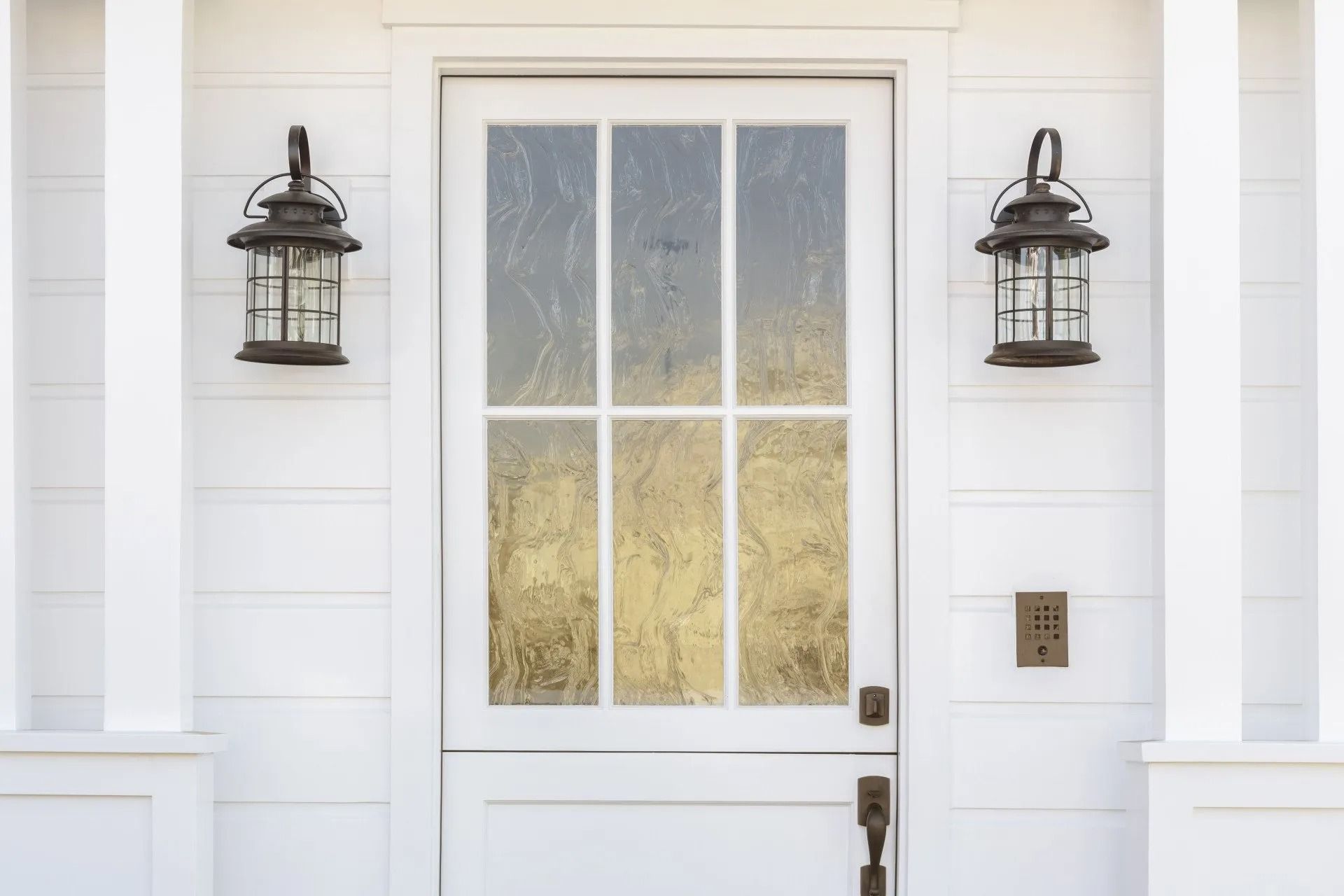 White door with six-pane window and two black lanterns on a white house exterior.