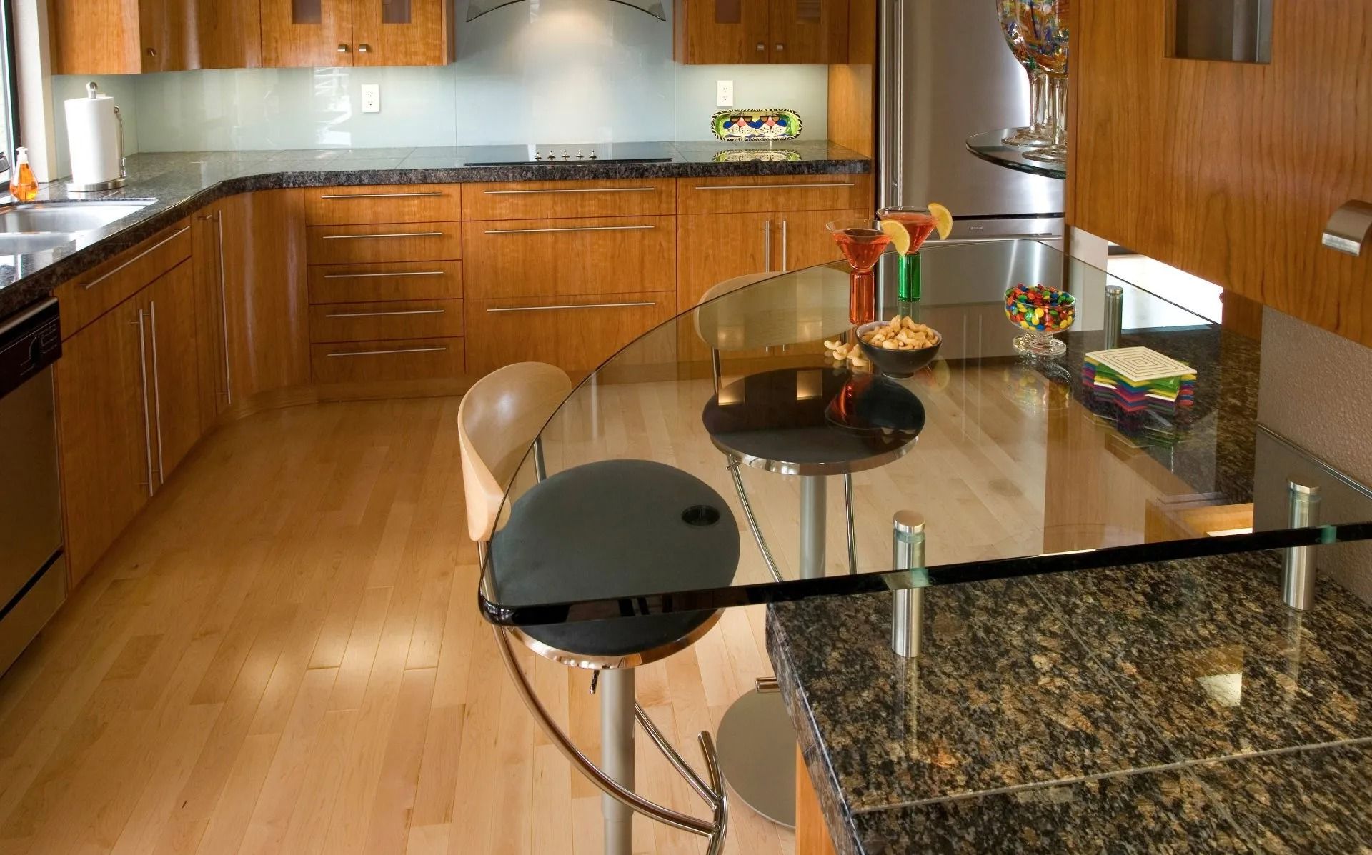 Modern kitchen with wood cabinets, granite countertops, and a glass breakfast bar.  Light wood floors and stainless steel appliances are also visible.