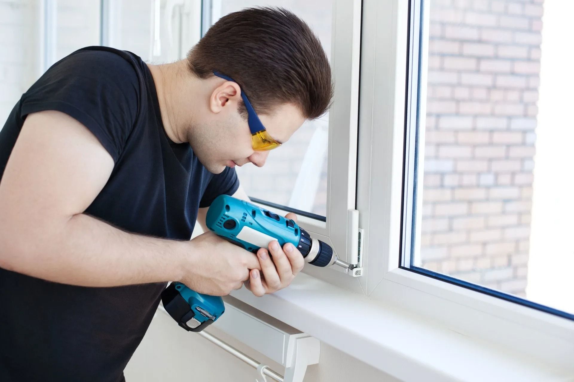 A man in a black shirt and safety glasses uses a blue cordless drill to work on a white window frame indoors.