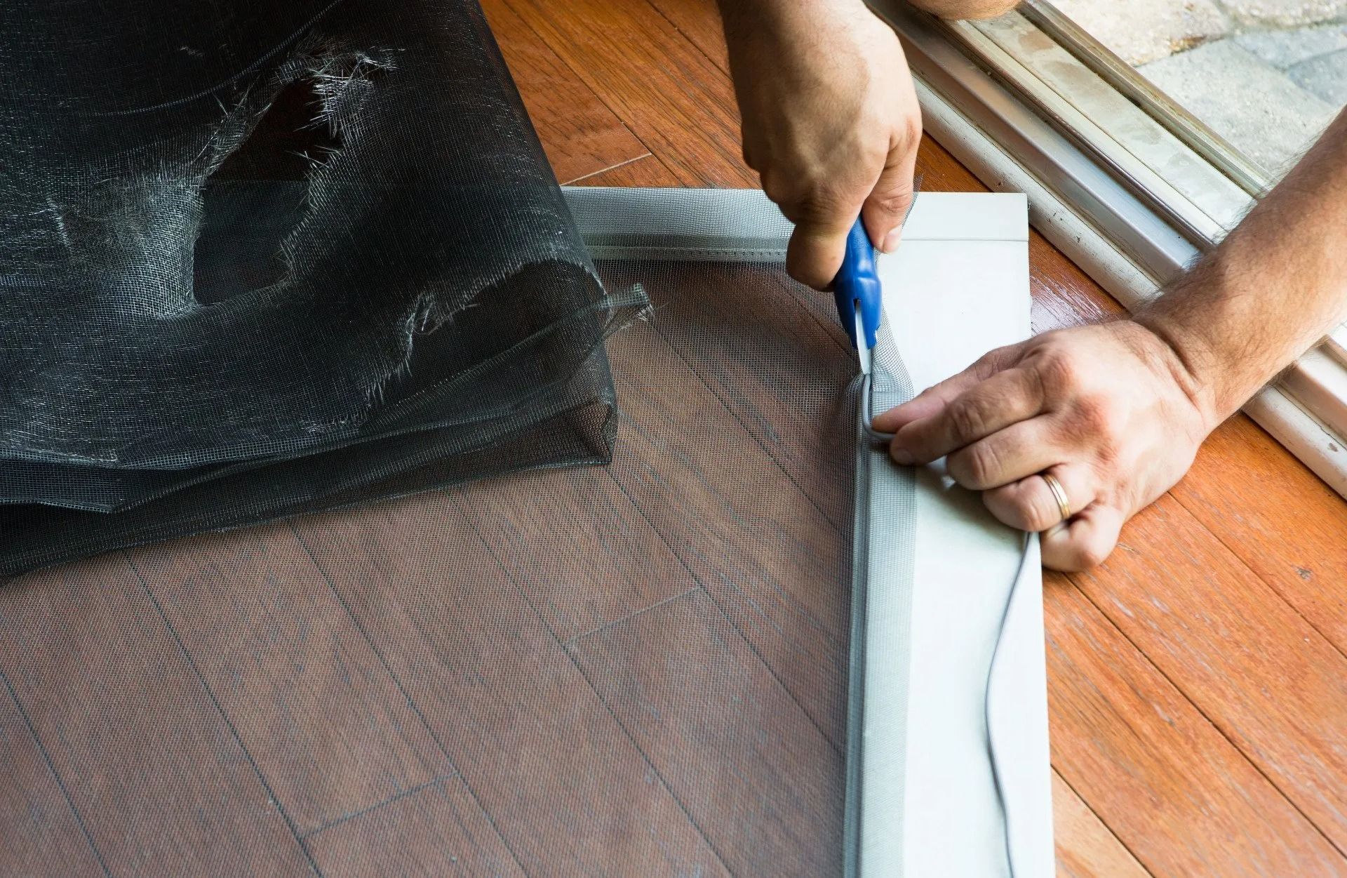 Person cutting excess screen material from a window screen frame with a utility knife, on a wooden floor.