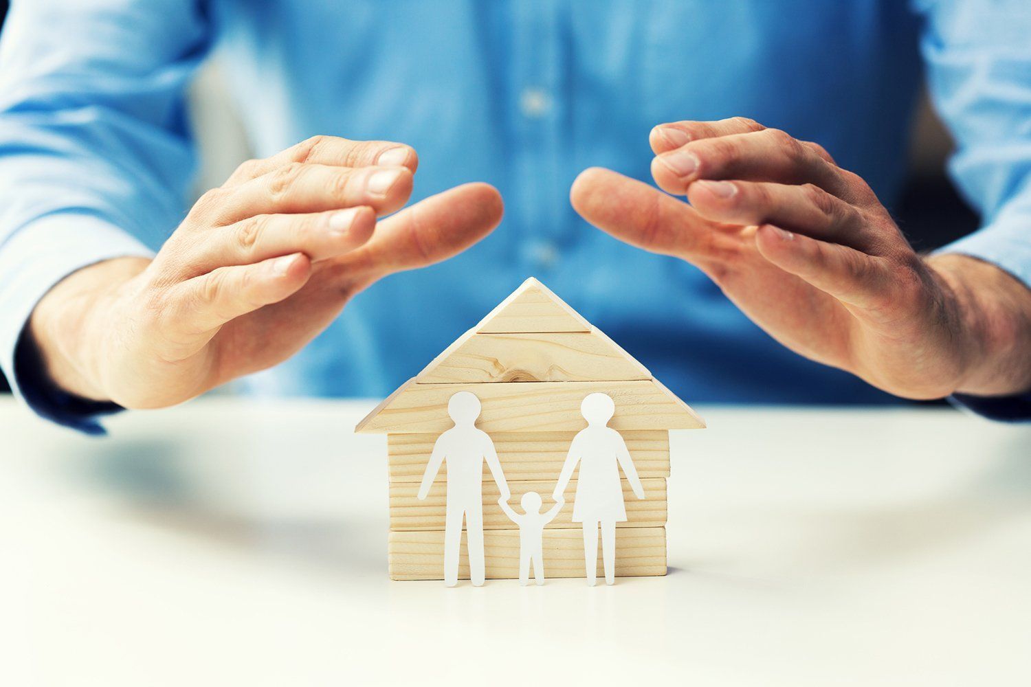 Hands protecting a paper house and family figures on a white surface