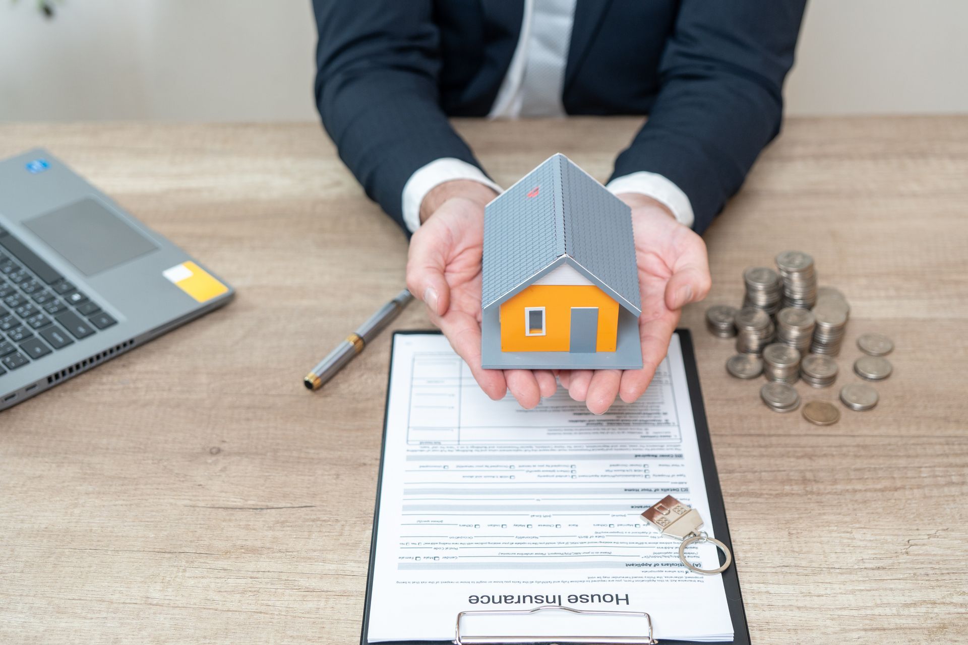 Hands holding a small house model over a mortgage application with laptop and coins on a desk
