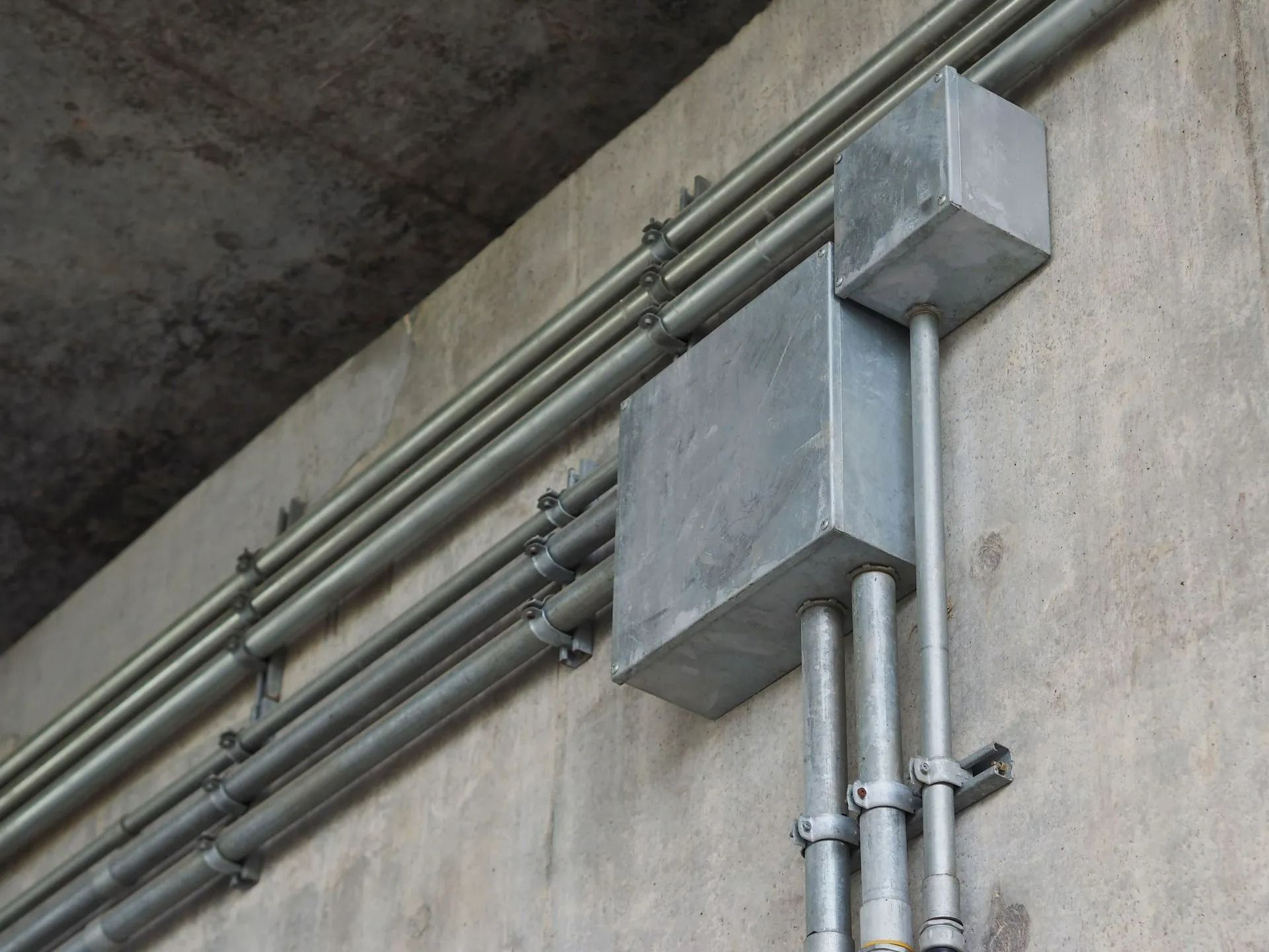 Gray electrical conduit and junction boxes mounted on a concrete wall near the ceiling.