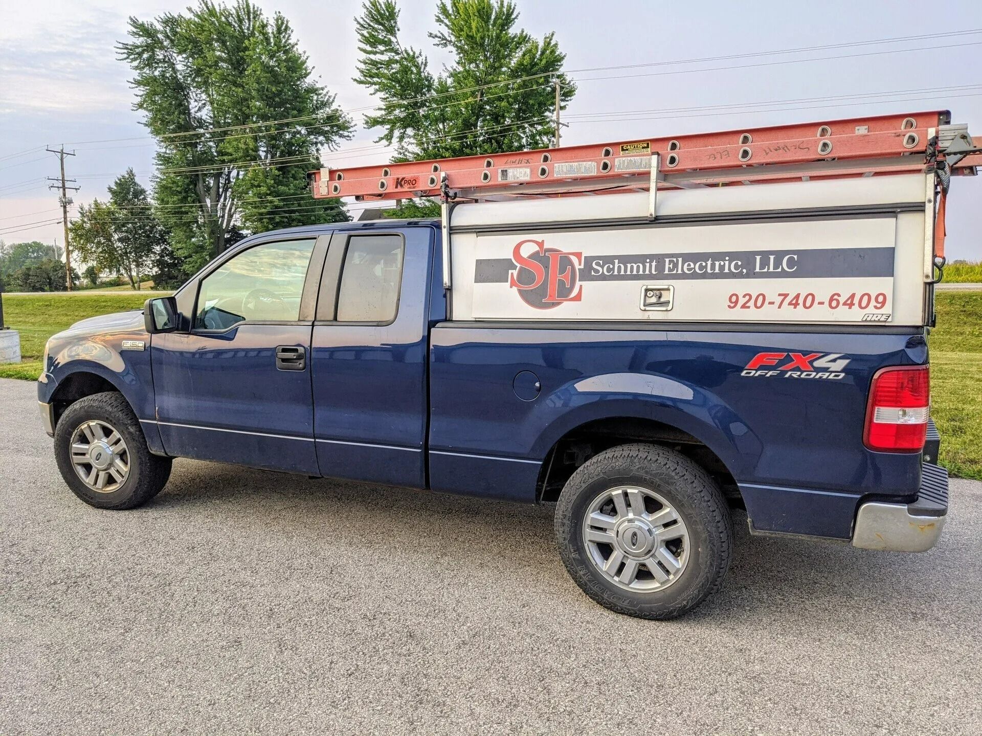 Blue utility pickup truck with ladder rack and business sign parked on gravel roadside.