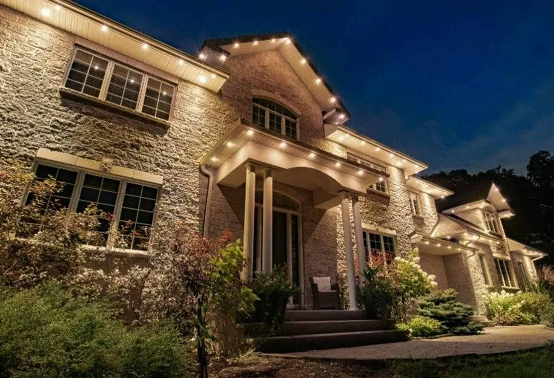Elegant brick house at dusk, warmly lit with string lights and a front porch surrounded by greenery