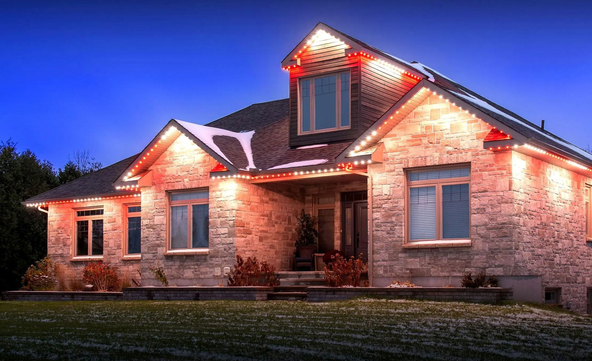 Stone house with warm exterior lights at dusk, featuring a lit front entry and dormer windows.