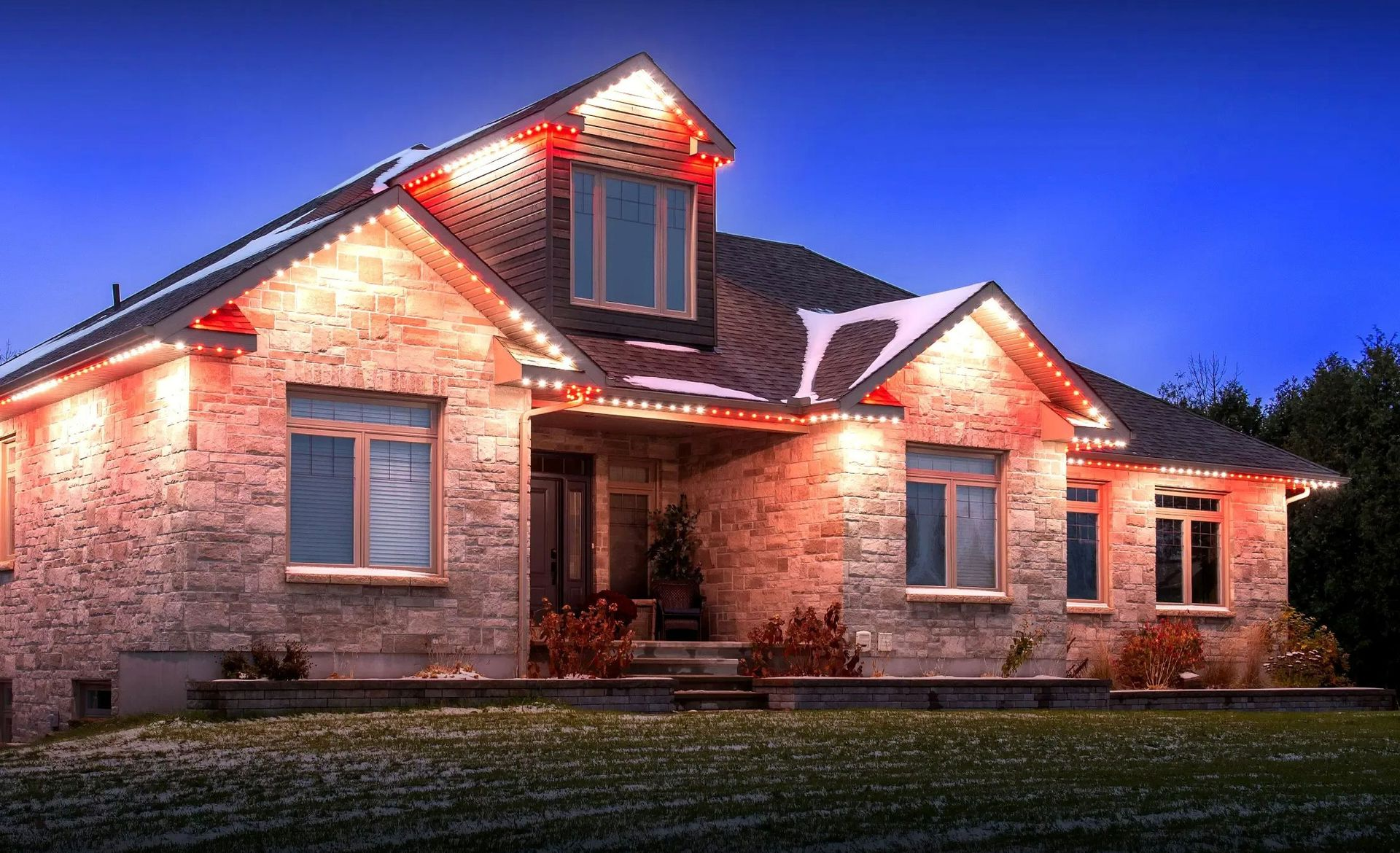 Stone house with lit Christmas lights at dusk, glowing against a blue evening sky