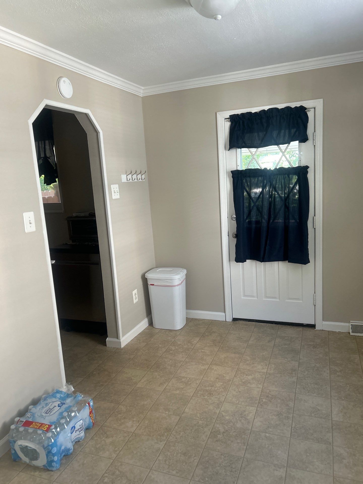 Small beige room with tiled floor, white door covered by a dark curtain, trash can, and baby items on the floor.