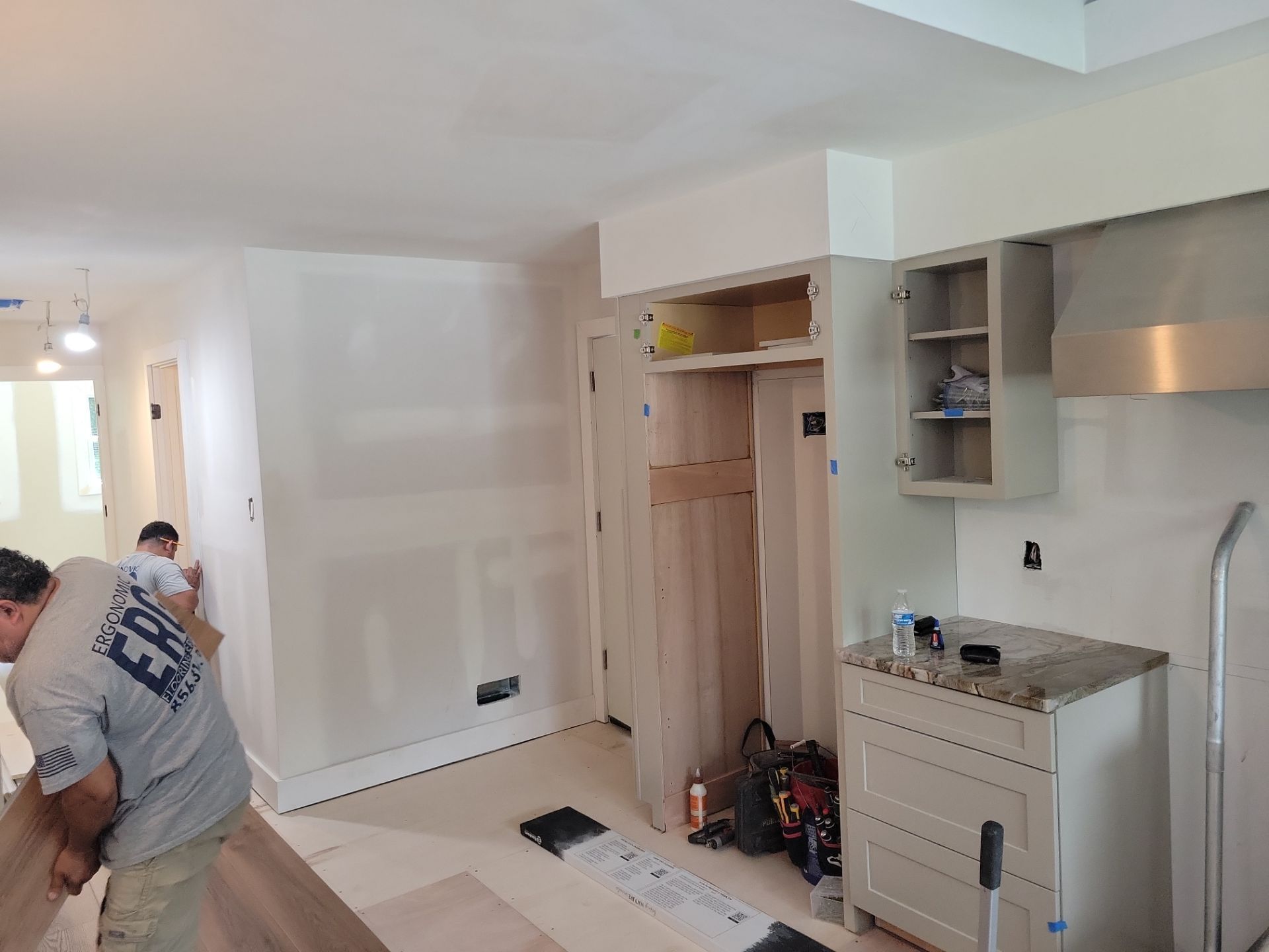Partially renovated kitchen with cabinets open and two workers installing flooring.