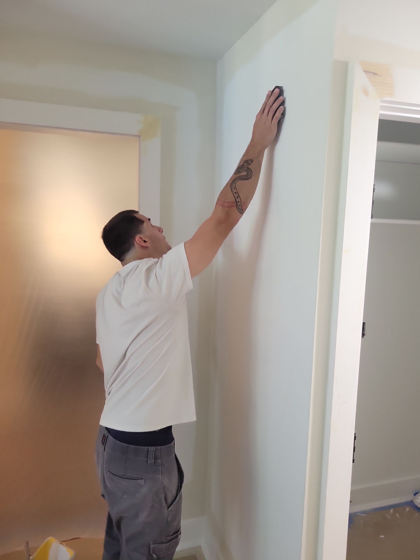Man in a white shirt reaching up to inspect a wall near a doorway in a hallway.