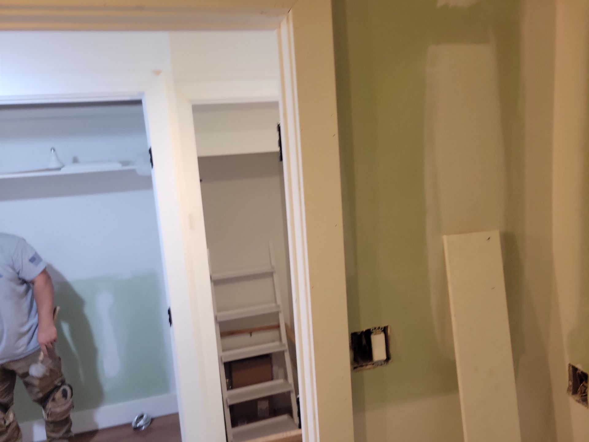 Partially painted room with a doorway, closet shelves, and a worker in a light blue shirt on the left.