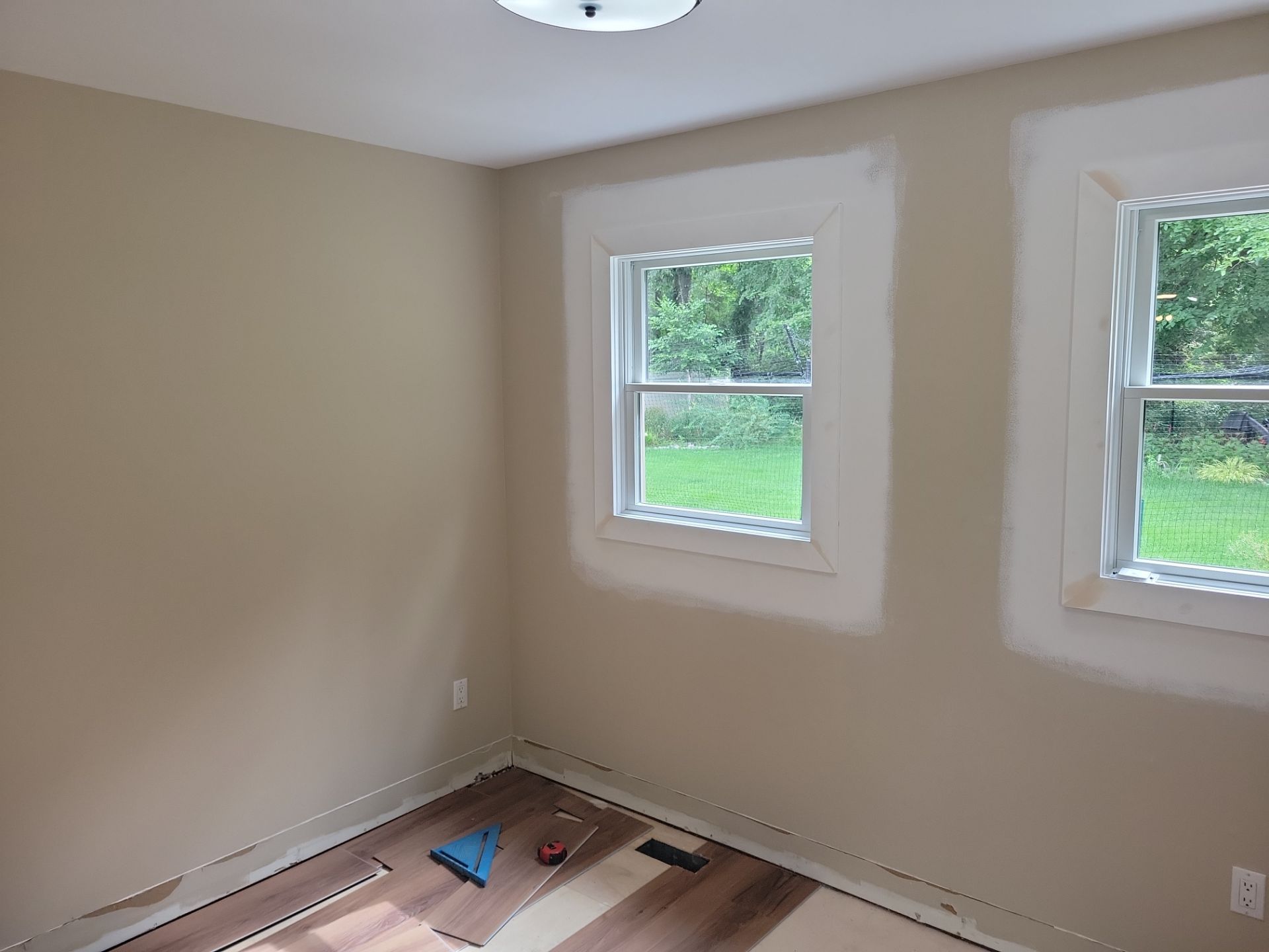 Empty beige room with two windows and unfinished flooring, paint supplies in the corner.