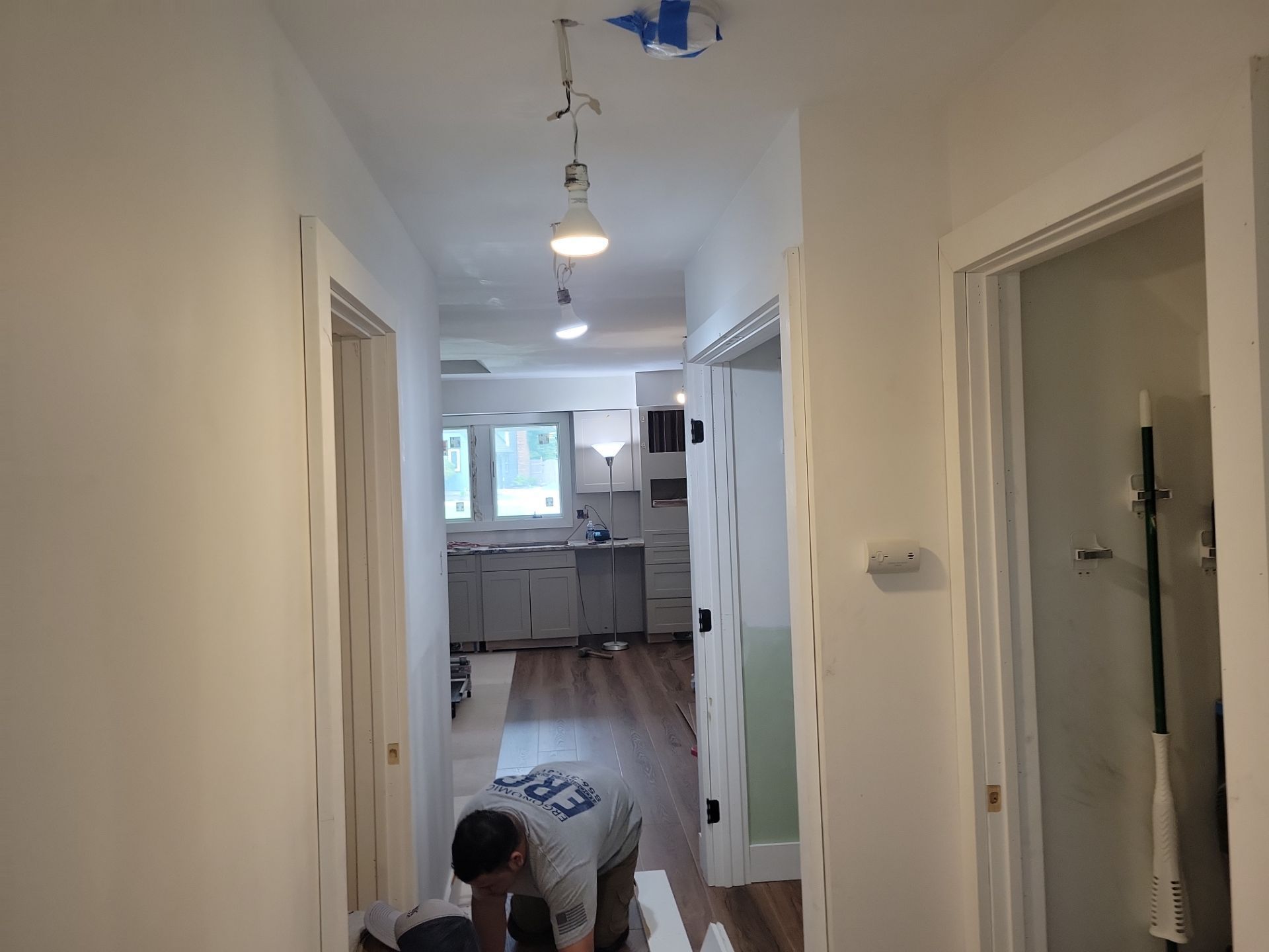 Narrow white hallway leading to a kitchen, with a person kneeling on the floor near the doorway.