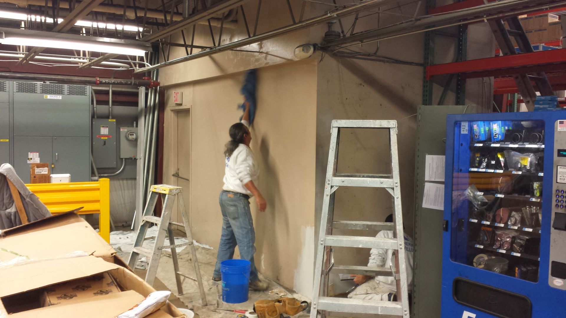 Person painting a beige wall in an industrial workshop, with ladders and equipment nearby.