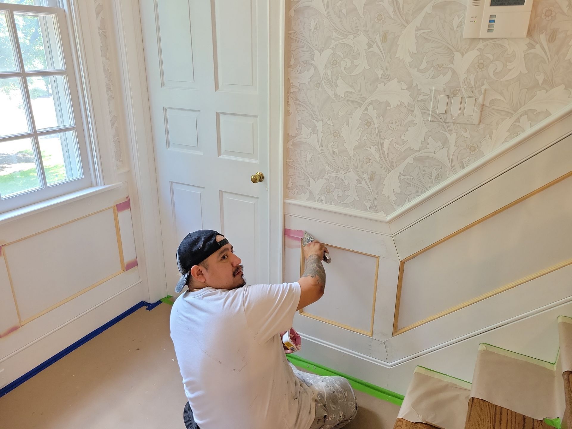 Painter working on a white stairwell wall with green tape and a window nearby.