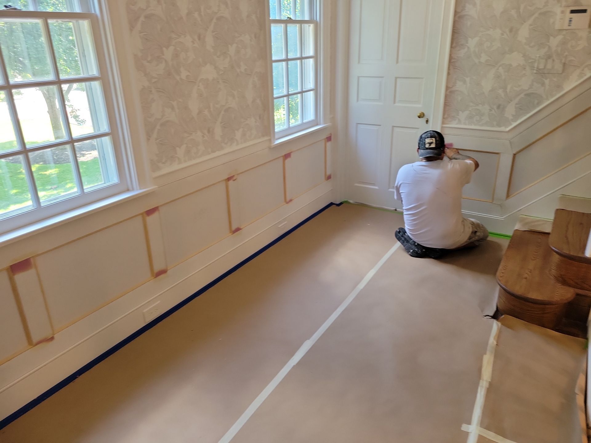 Person crouching on the floor in a room with taped-off flooring, windows, and a closed door.