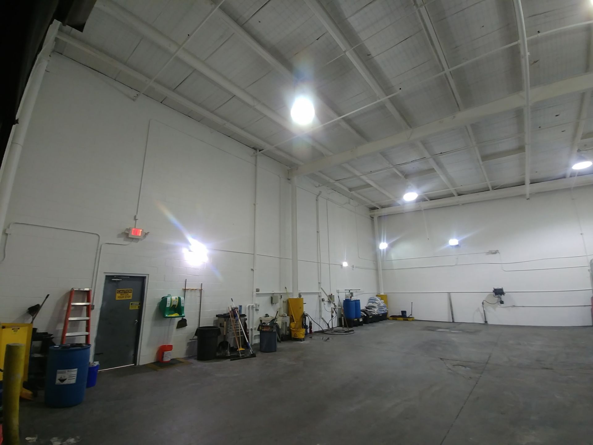 Empty warehouse interior with high white ceiling, gray floor, and equipment lined along the wall.