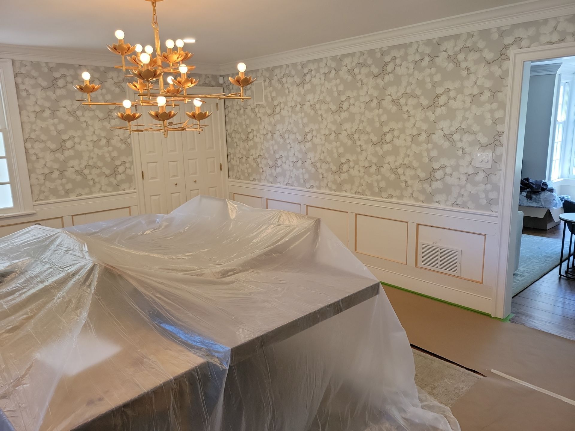 Dining room with chandelier, patterned wallpaper, and furniture covered in plastic sheeting during renovation.