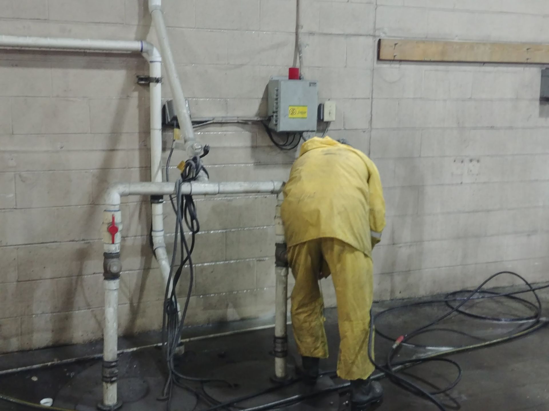 Worker in yellow protective gear cleaning a wet industrial floor with a hose near wall-mounted equipment.