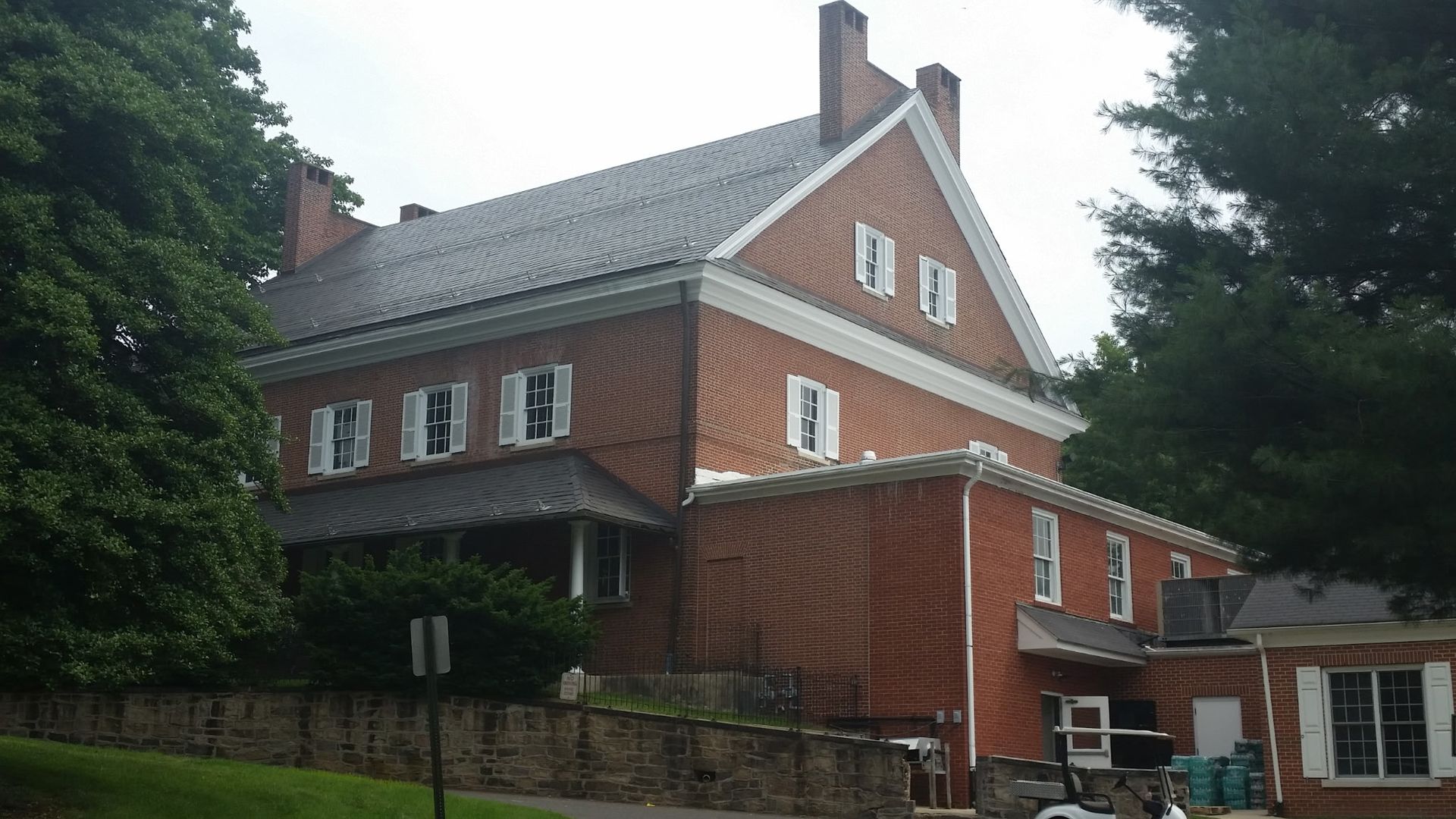 Large red-brick house with white trim and a steep roof, surrounded by trees and a stone wall.